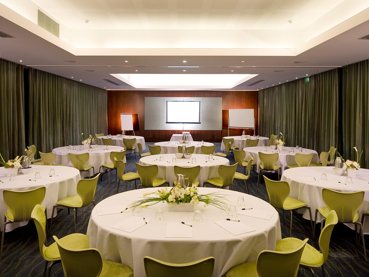 A Meeting Room With Round Tables, Green Chairs And A Tv at The Front Of The Room.