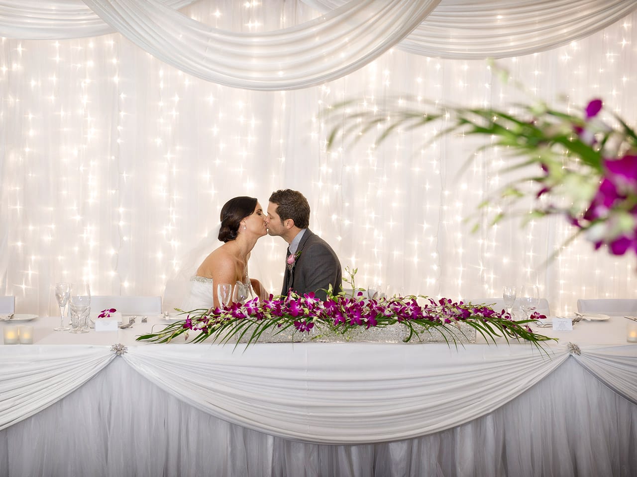 A Wedding Table With The Groom And Bride Sitting And Kissing.