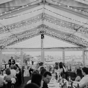 Black And White Capture of Wedding Reception Inside the Function Room With string Lights With Bride And Groom Sitting In Front , A Speaker And Guests