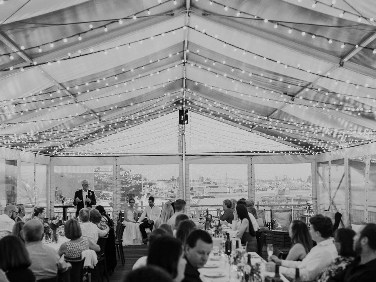 Black And White Capture of Wedding Reception Inside the Function Room With string Lights With Bride And Groom Sitting In Front , A Speaker And Guests