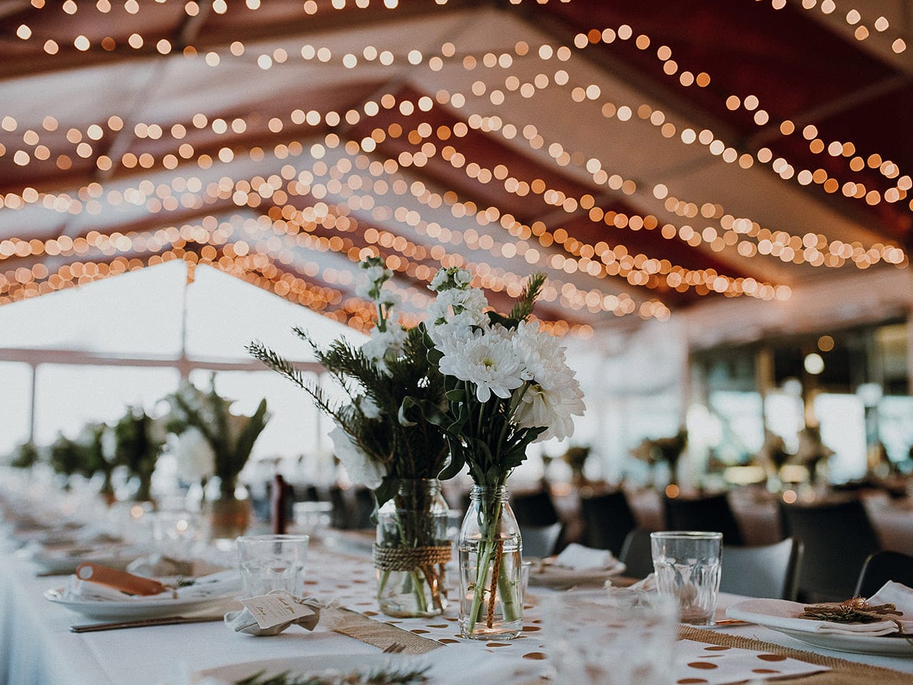 Inside the Function Room with Flower Centerpieces in Long Tables, String Lights and Drapings on the Ceiling