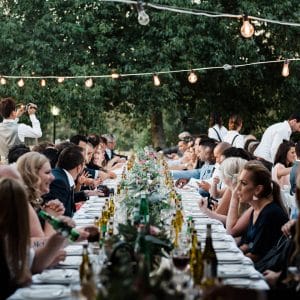 People Gathered In A Long Table With Food And Drinks And A String Lights In The Terrace