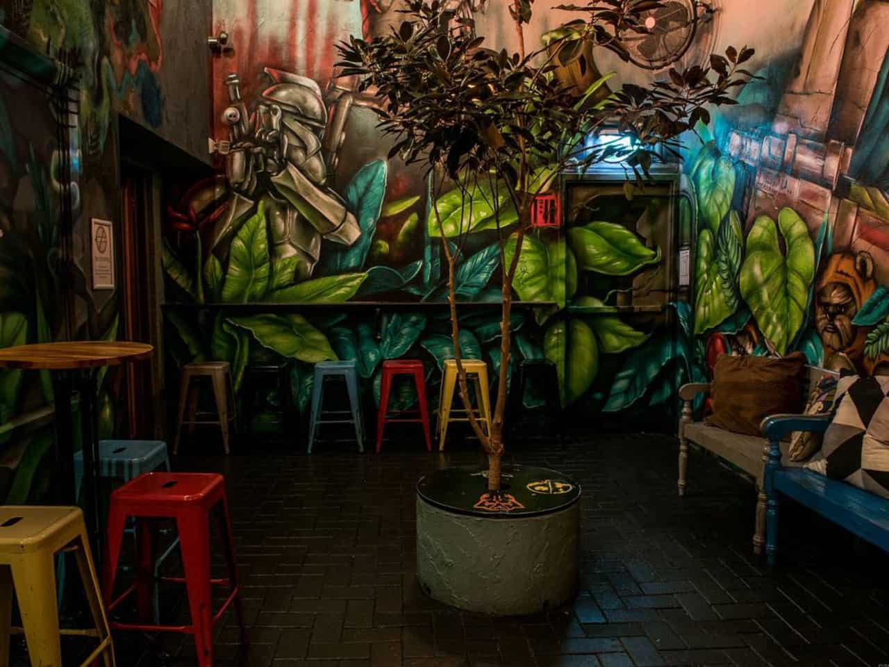The Courtyard With Side Tables And Colorful High Chairs, Painted Walls, Pillows And A Small Tree