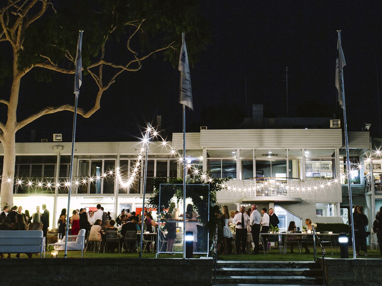 People Gathering Outside The Function Area Under A Tree With Tables, Chairs, String Lights And Flag Poles