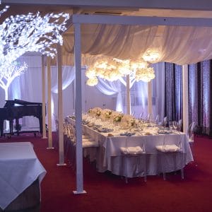 A Long Table Setup With Flower Centerpieces, Flower Chandelier, Ceiling Drapping, Piano And Artificial Tree Lights