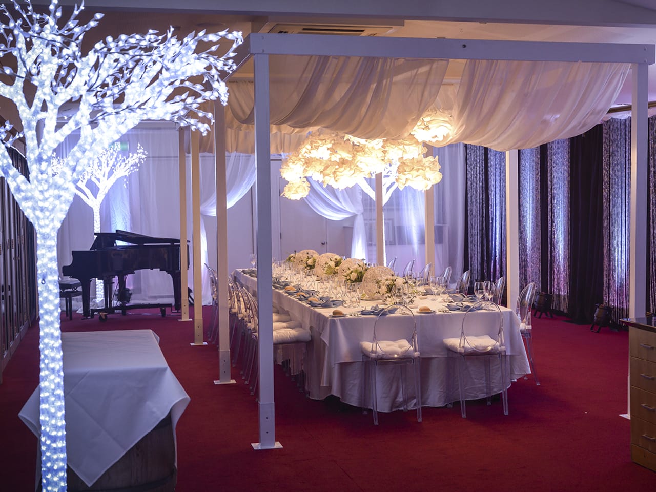 A Long Table Setup With Flower Centerpieces, Flower Chandelier, Ceiling Drapping, Piano And Artificial Tree Lights