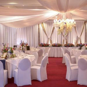 Tables And Chairs In Banquet Style And A Special Long Table With Flower Centerpieces, Chandelier And Ceiling Drapping