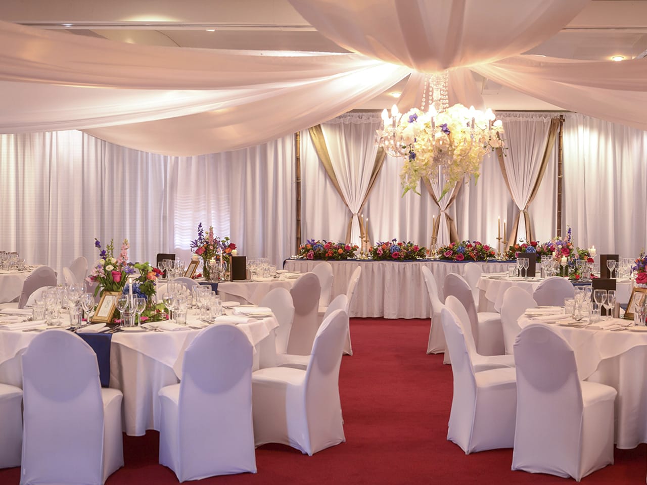 Tables And Chairs In Banquet Style And A Special Long Table With Flower Centerpieces, Chandelier And Ceiling Drapping