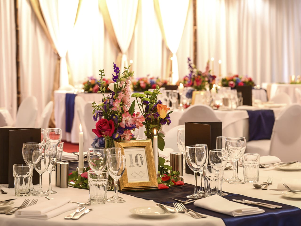 Flower Centerpieces With Table Number, Wine Glasses, Table Napkins, Spoon And Forks And Stage Behind