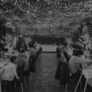 Guests Seated In Long Tables In The Open Air Function Space With String Lights Above Them