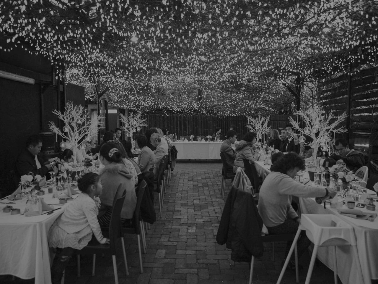 Guests Seated In Long Tables In The Open Air Function Space With String Lights Above Them