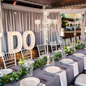 Chairs And A Long Table Setup With Green Leafy Centerpieces And 'I Do' Standee Inside The Function Room