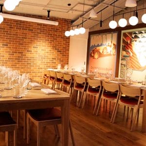 Two Long Tables With Chairs Inside The Function Room With Brick Walls And Warm Lighting