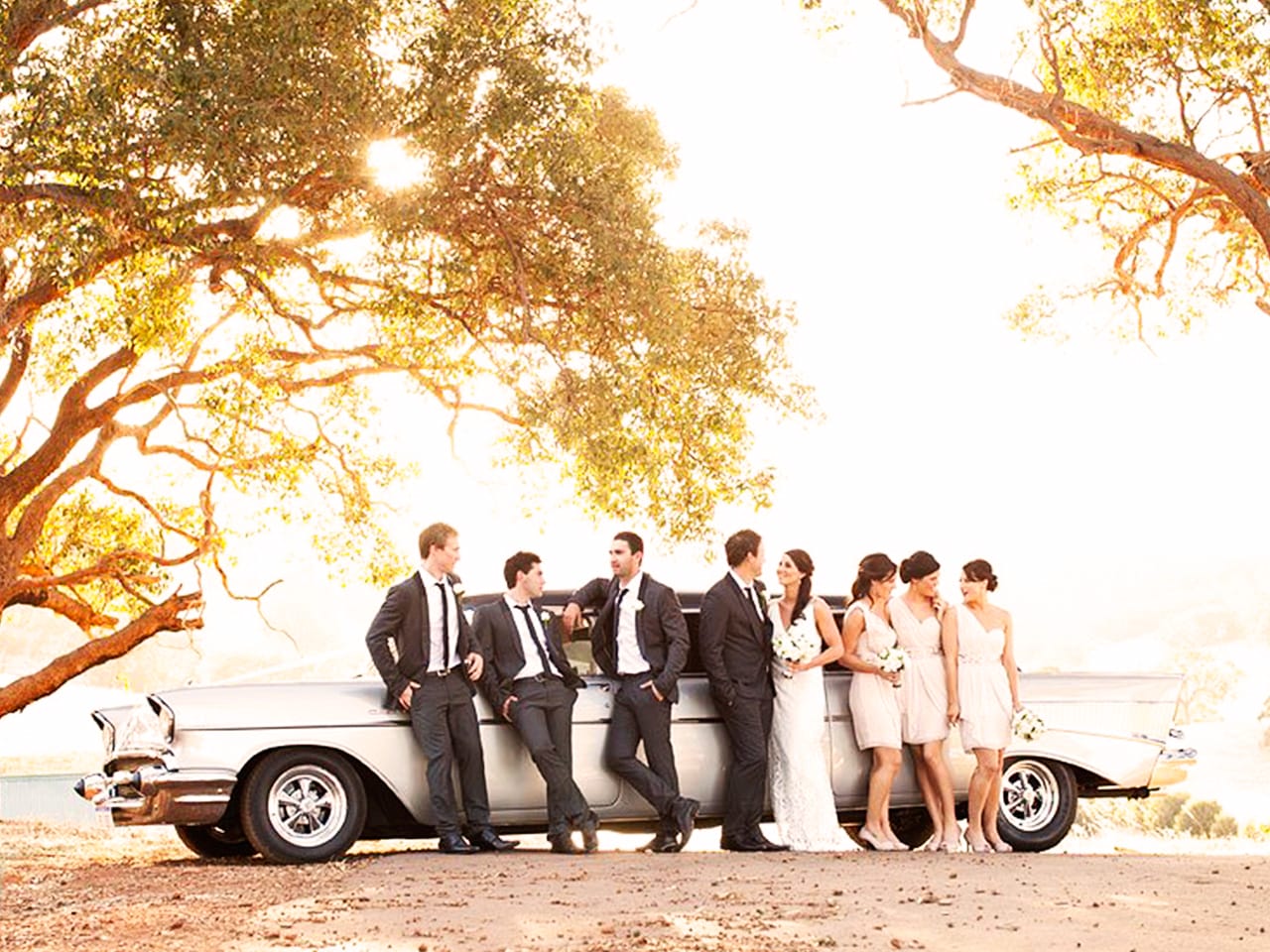 Bride And Groom With The Entourage Taking Photo Under The Trees With A Vintage Car Behind Them