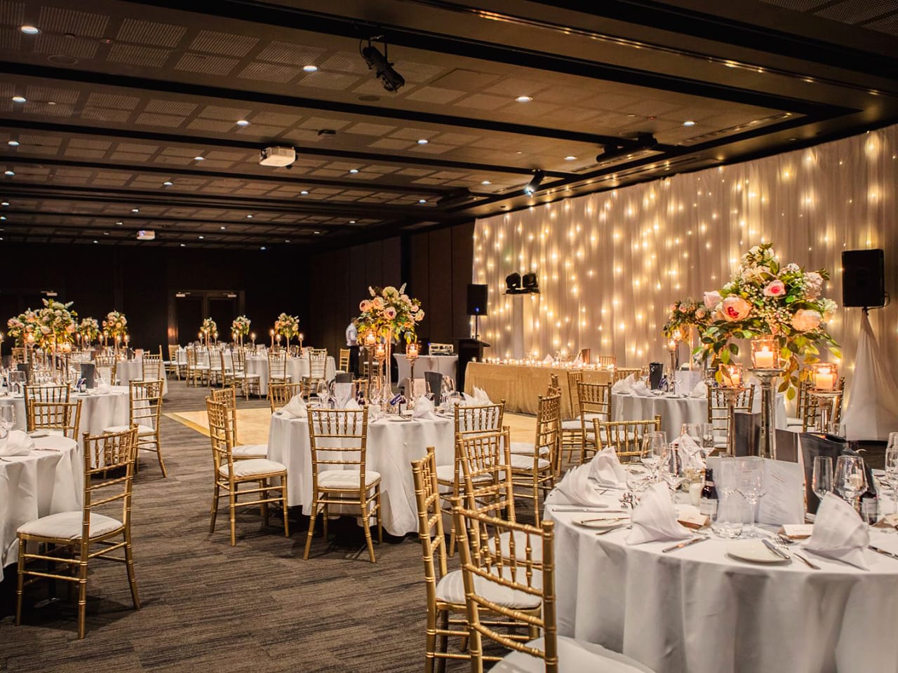 Tables And Chairs In Banquet Style With Flower Centerpieces And String Lights On The Front